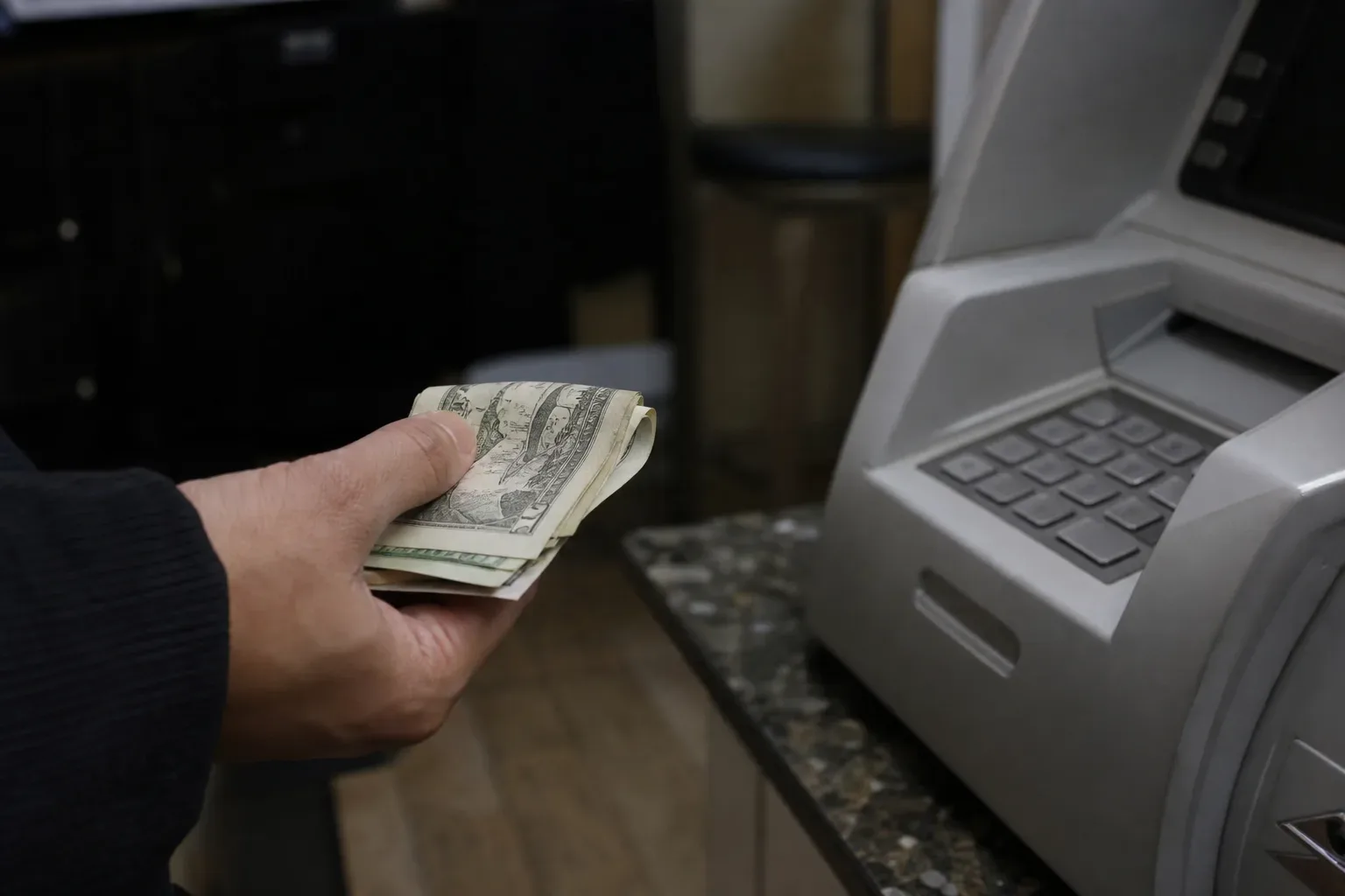 A hand holding several folded cash bills next to a small countertop ATM machine