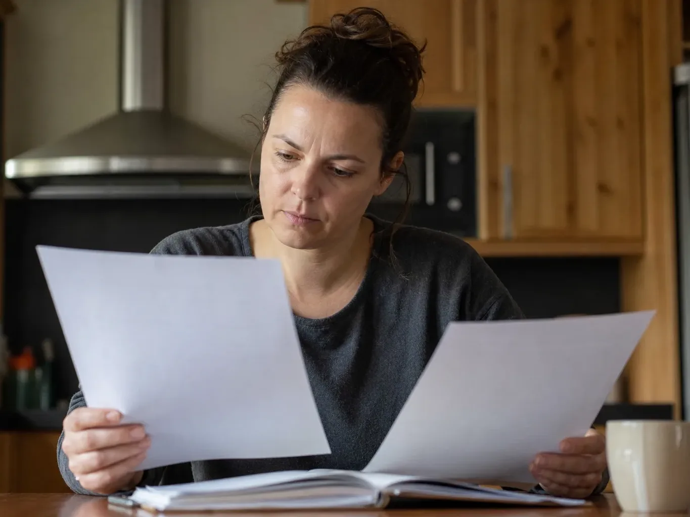 A person sitting at a kitchen table comparing two documents with a thoughtful expression